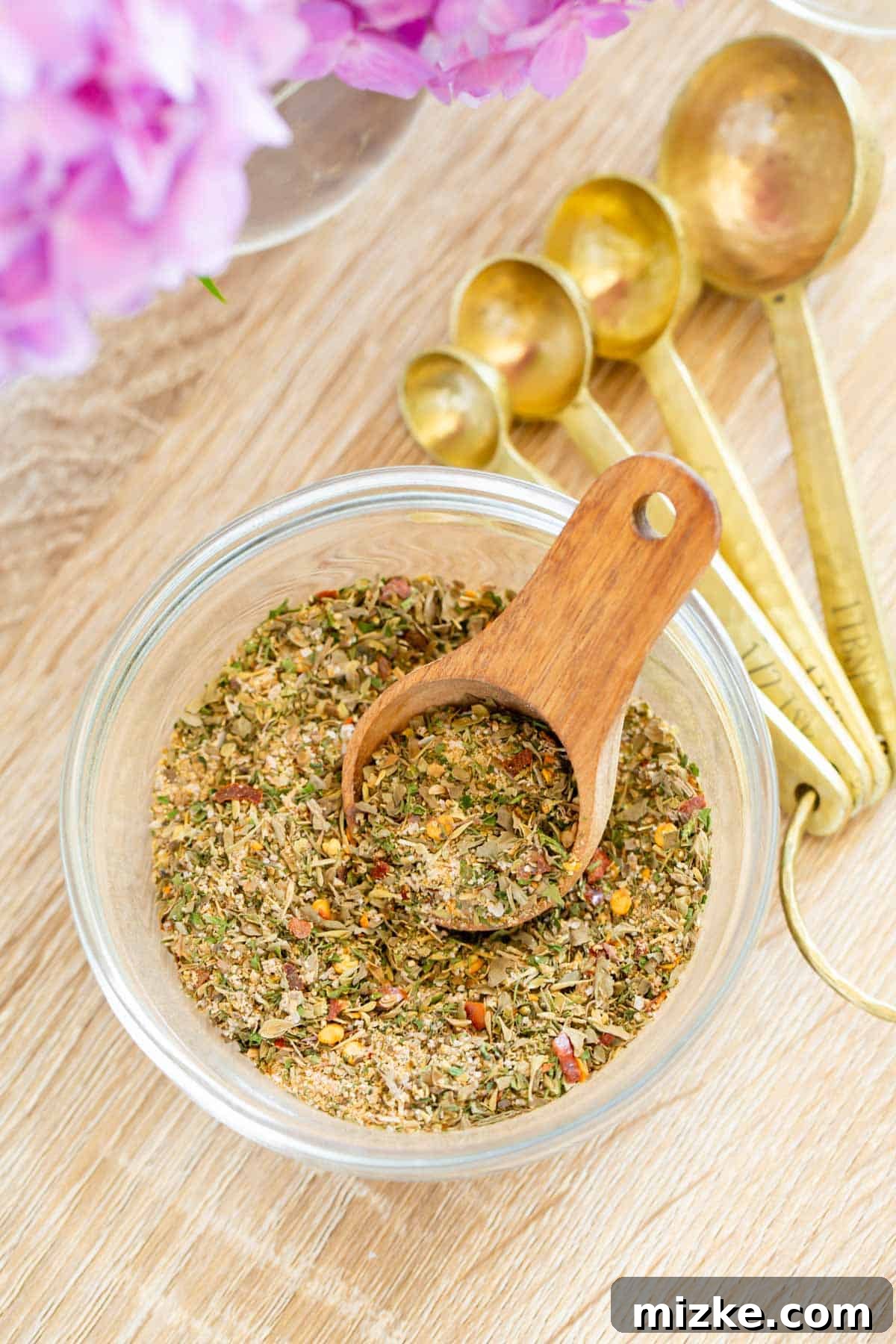 Aegean Sun Seasoning 3 greek seasoning in a clear bowl with a small wooden spoon next to gold measuring spoons