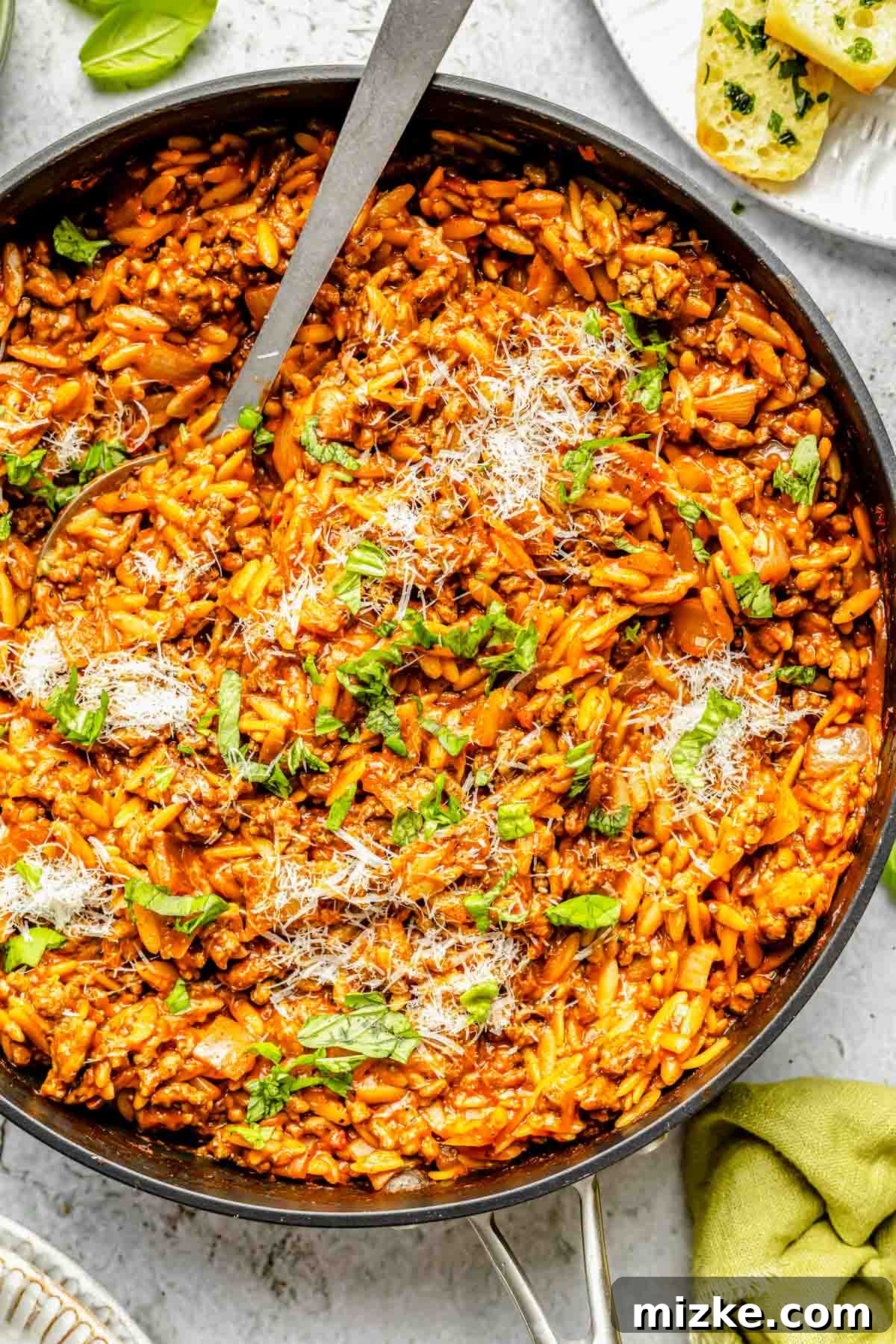 Hearty Beef and Orzo 2 A vibrant, steaming skillet of ground beef and orzo pasta, richly coated in a creamy tomato sauce. It's elegantly garnished with fresh basil leaves and a generous sprinkle of grated Parmesan cheese, with a metal spoon resting in the dish, ready for serving.