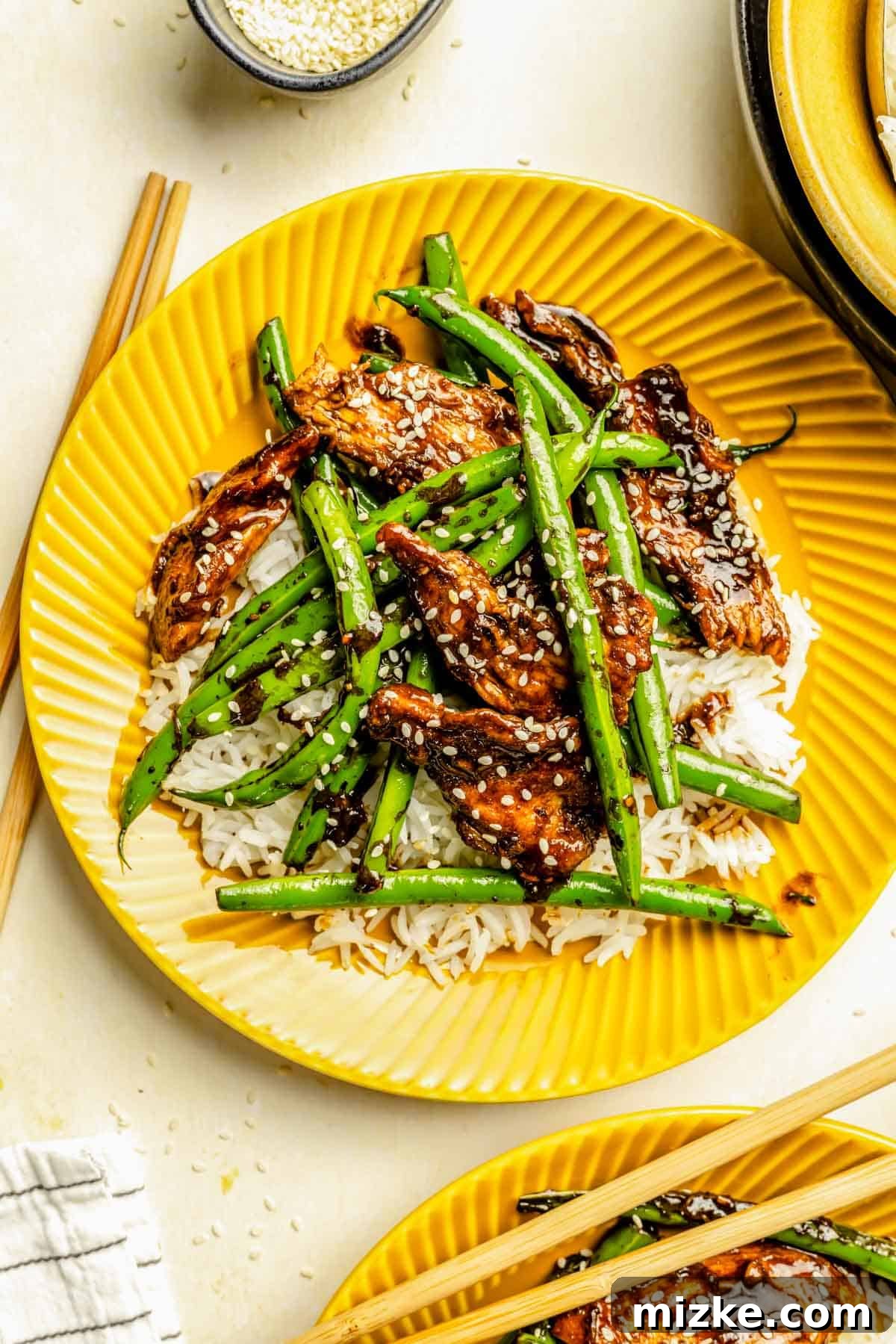 Chicken Stir Fry with Crisp Green Beans 2 Green beans and chicken stir fry served over white rice on a yellow plate, garnished with white sesame seeds.