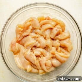 Raw thin slices of chicken marinating in a clear bowl, coated in cornstarch, soy sauce, baking soda, and water.