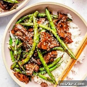 A pink bowl of green bean beef stir fry with white rice and wooden chopsticks