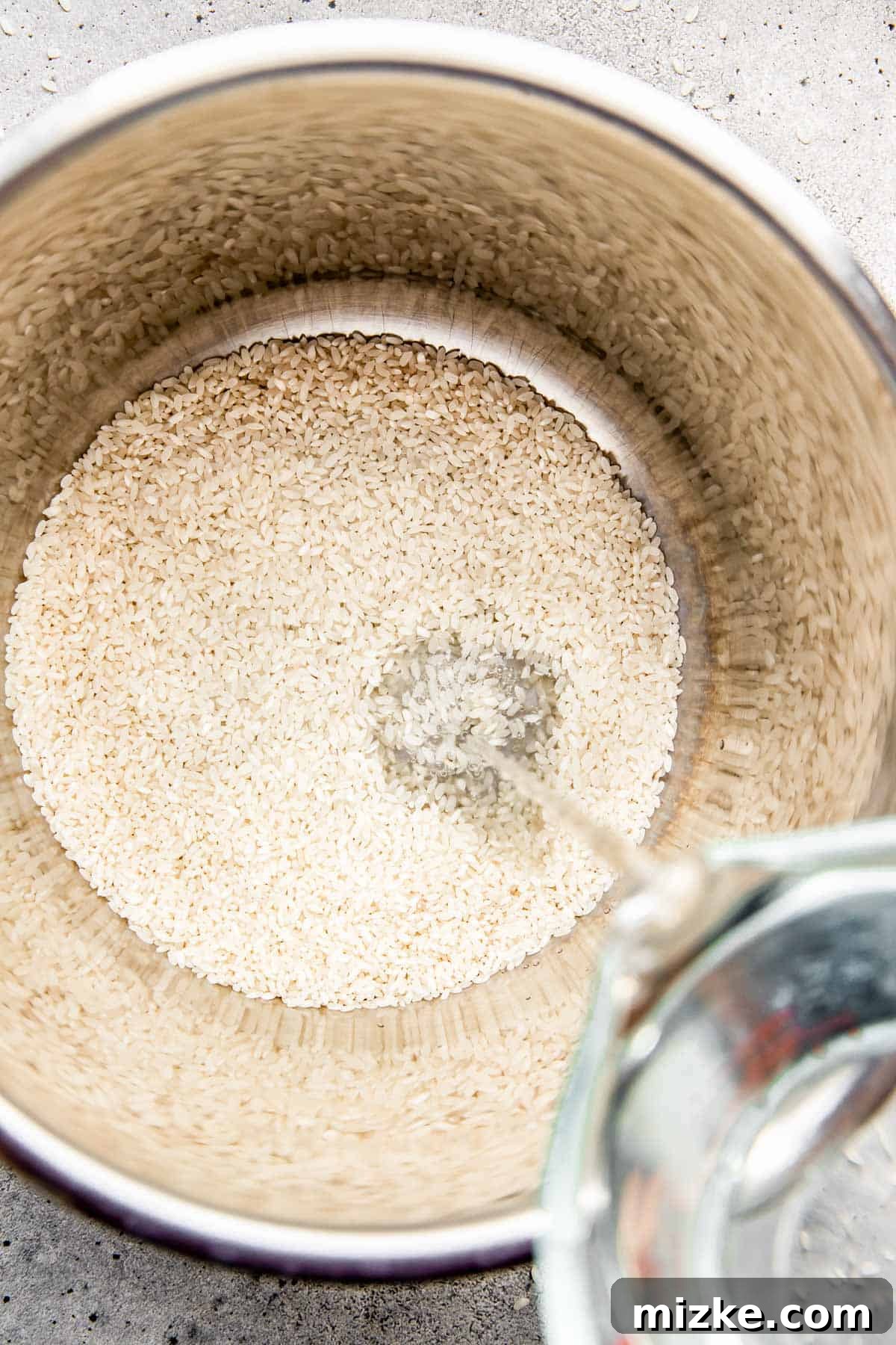The Secret to Perfectly Fluffy White Rice 3 White rice grains are shown in a stainless steel pot as cold water is carefully poured over them, preparing for the cooking process.