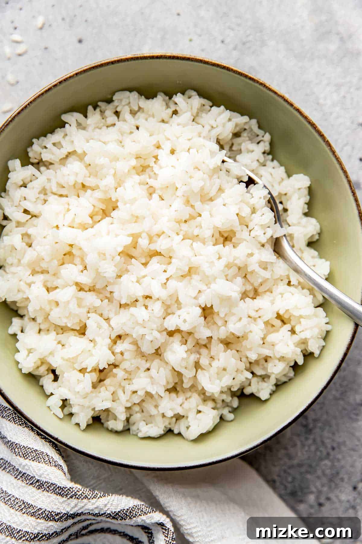 The Secret to Perfectly Fluffy White Rice 7 A vibrant green bowl brimming with beautifully cooked white rice, accompanied by a sleek metal spoon, all resting on a striped linen towel.