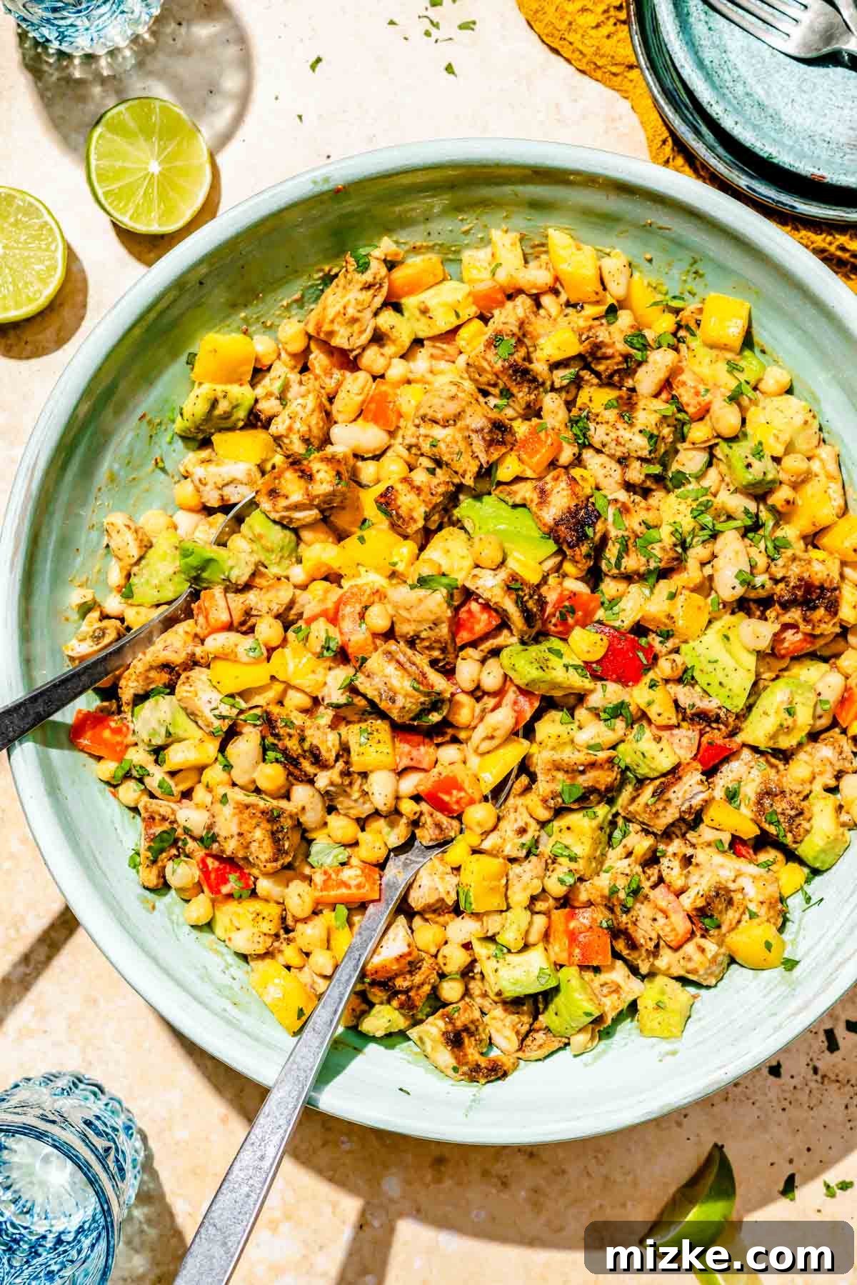 Chipotle Chicken Hearty Bean Salad 2 A large blue bowl filled with vibrant chipotle chicken dense bean salad, tossed with metal serving utensils. Fresh lime wedges and glasses of water are placed beside the bowl, highlighting the salad's refreshing and zesty character.