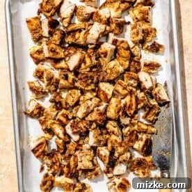Cubed grilled chipotle chicken thighs arranged neatly on a metal baking sheet lined with parchment paper, with a knife resting alongside, ready for assembly.