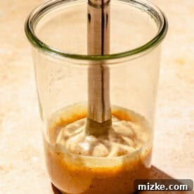 An immersion blender actively blending all the spicy chipotle dressing ingredients in a tall glass container, creating a perfectly smooth and creamy consistency.