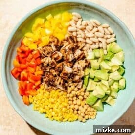 A large blue bowl showcasing a colorful array of diced red and yellow bell peppers, cubed chipotle chicken, great northern beans, cubed avocados, corn kernels, and chickpeas, all mixed together before the dressing is added.