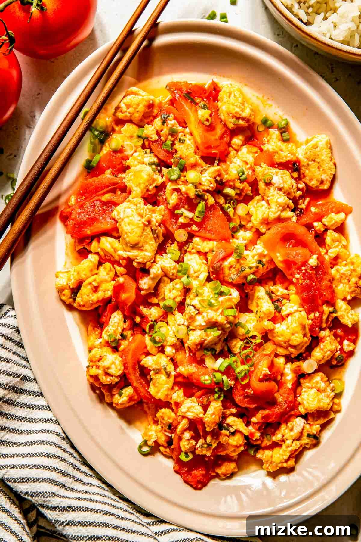 Savory Tomato Egg Scramble 2 A beautifully presented dish of fluffy scrambled eggs mixed with vibrant, tender tomato wedges and garnished with fresh chopped green onions. The inviting stir-fry is served on a modern beige oval plate, with elegant chopsticks resting alongside, ready for enjoyment.
