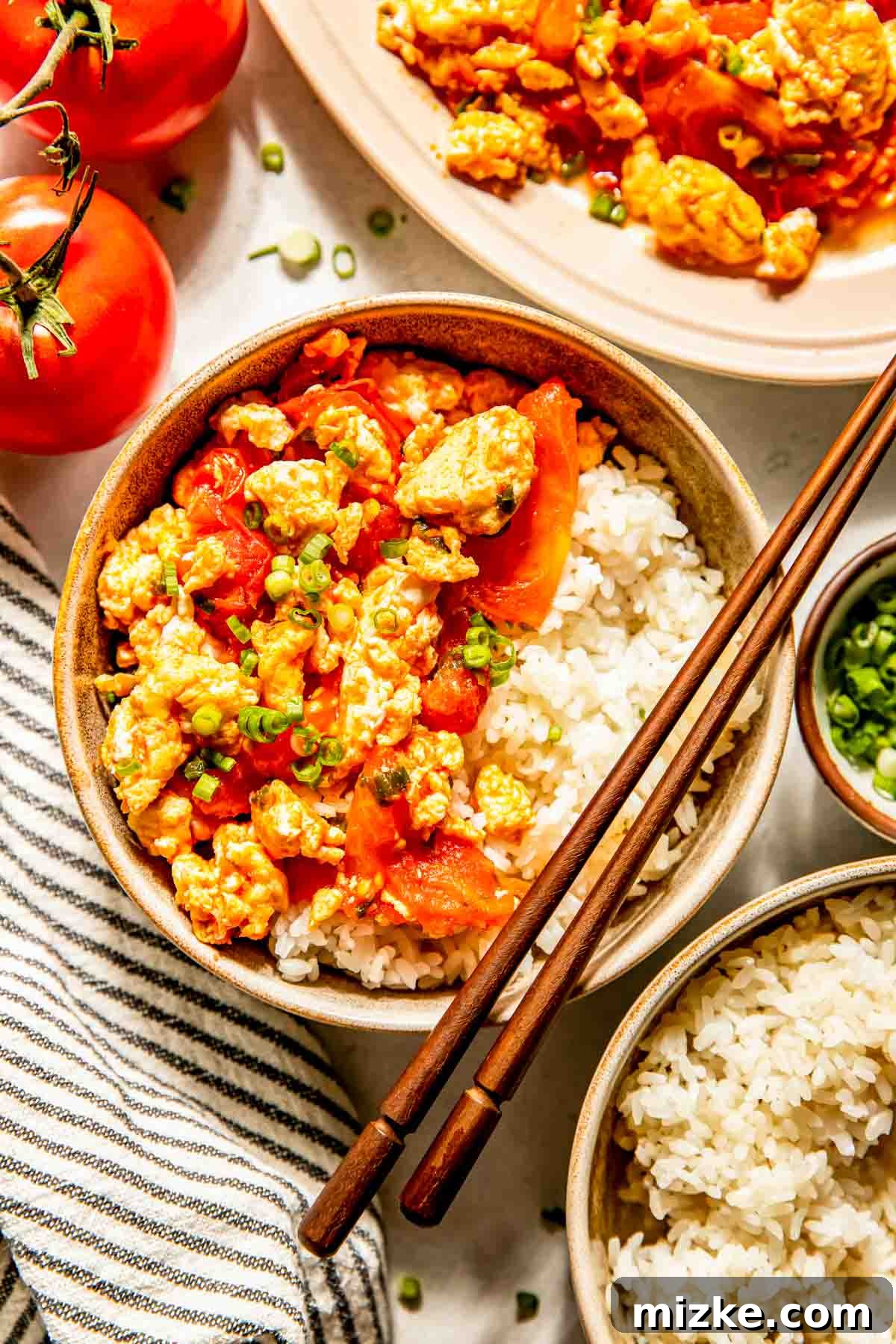 Savory Tomato Egg Scramble 3 A steaming bowl of fluffy white rice crowned with the savory and tangy Chinese tomato and egg stir fry, vibrantly garnished with chopped green onions. Whole ripe tomatoes and a stylish striped napkin sit beside the bowl, with chopsticks resting gracefully across it, inviting you to dig in.