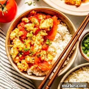 Bowl of rice topped with scrambled eggs and tomatoes, garnished with green onions, next to whole tomatoes and a striped napkin. Chopsticks rest on the bowl.