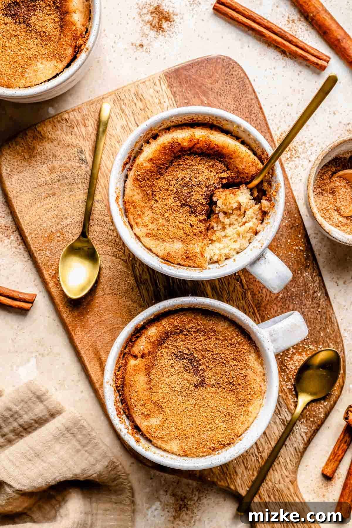 Snickerdoodle Mug Magic 3 Two mugs of snickerdoodle mug cake on a wooden board with spoons and cinnamon sticks nearby.