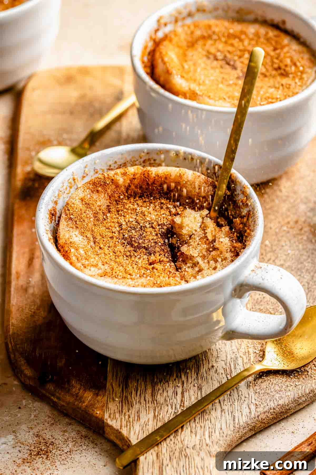 Snickerdoodle Mug Magic 4 Two white ceramic cups filled with snickerdoodle mug cake, cinnamon sugar-topped, with spoons. The cups are placed on a wooden board with an additional gold spoon beside them.