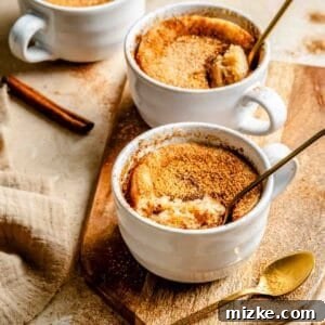 Three white mugs filled with snickerdoodle mug cakes on a wooden board, accompanied by gold spoons and cinnamon sticks.