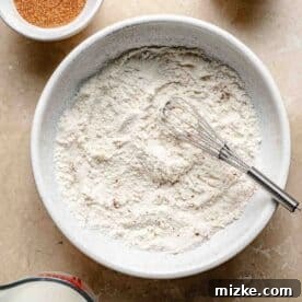Snickerdoodle Mug Magic 6 A bowl of flour with a whisk inside is surrounded by small bowls and a measuring cup containing cinnamon, vanilla, melted butter, and milk on a beige countertop.