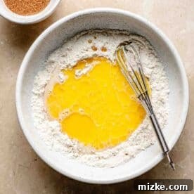 Snickerdoodle Mug Magic 7 Bowl of flour with melted butter and a whisk. A small bowl of cinnamon sugar is next to it on a beige surface.