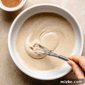Snickerdoodle Mug Magic 8 Person whisking batter in a bowl next to a small bowl of cinnamon sugar on a marble surface.