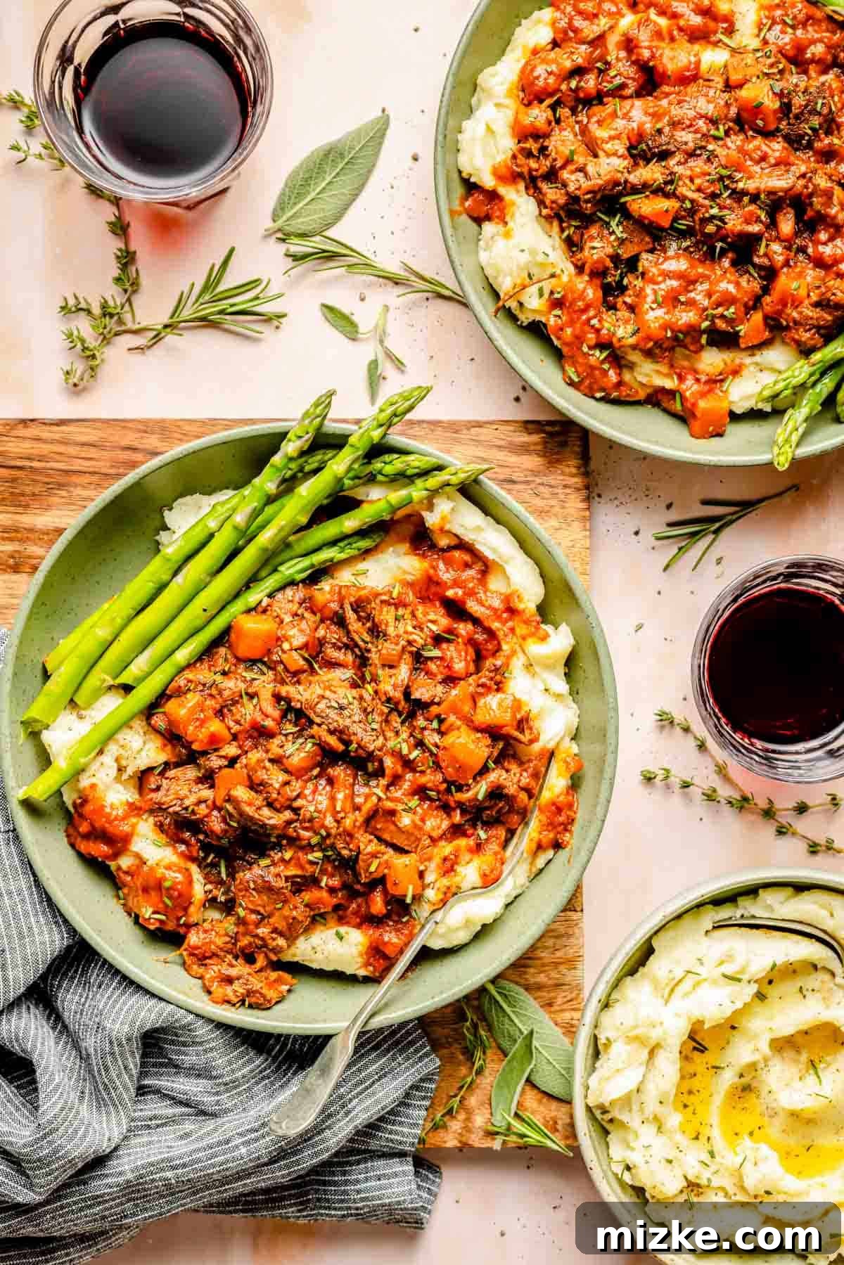Slow-Braised Lamb Ragu 2 Two plates of fluffy mashed potatoes generously topped with rich, slow-braised lamb ragu and a side of vibrant green asparagus. The setting includes two glasses of deep red wine and a bowl of extra mashed potatoes, with fresh herb sprigs scattered for garnish, creating an inviting and elegant dining scene.
