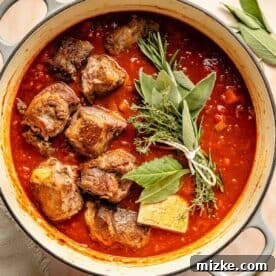 A pot of braised lamb meat in tomato sauce garnished with fresh herbs, including rosemary, thyme, and bay leaves.