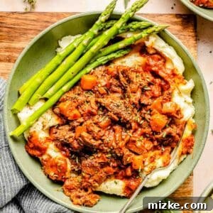 A plate of mashed potatoes topped with lamb ragu and served with asparagus. A napkin and herbs are on the table beside the dish.