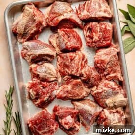 Raw, seasoned lamb shoulder pieces are arranged on a metal tray. Fresh herbs, including rosemary and thyme, are placed beside the tray.