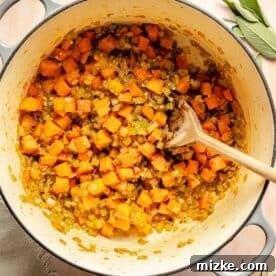 Chopped vegetables, including carrots, onions, and celery, cooked in a large pot with herbs and a wooden spoon.