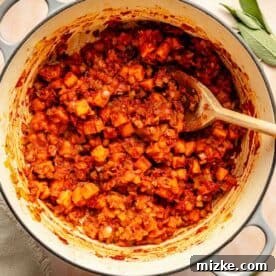 A pot of diced vegetables in a red sauce. A wooden spoon is immersed in the mixture. Fresh herbs are scattered around the pot.