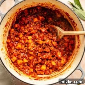 A pot of vegetables in a rich red sauce, accompanied by a wooden spoon. Fresh herbs are scattered around the pot.