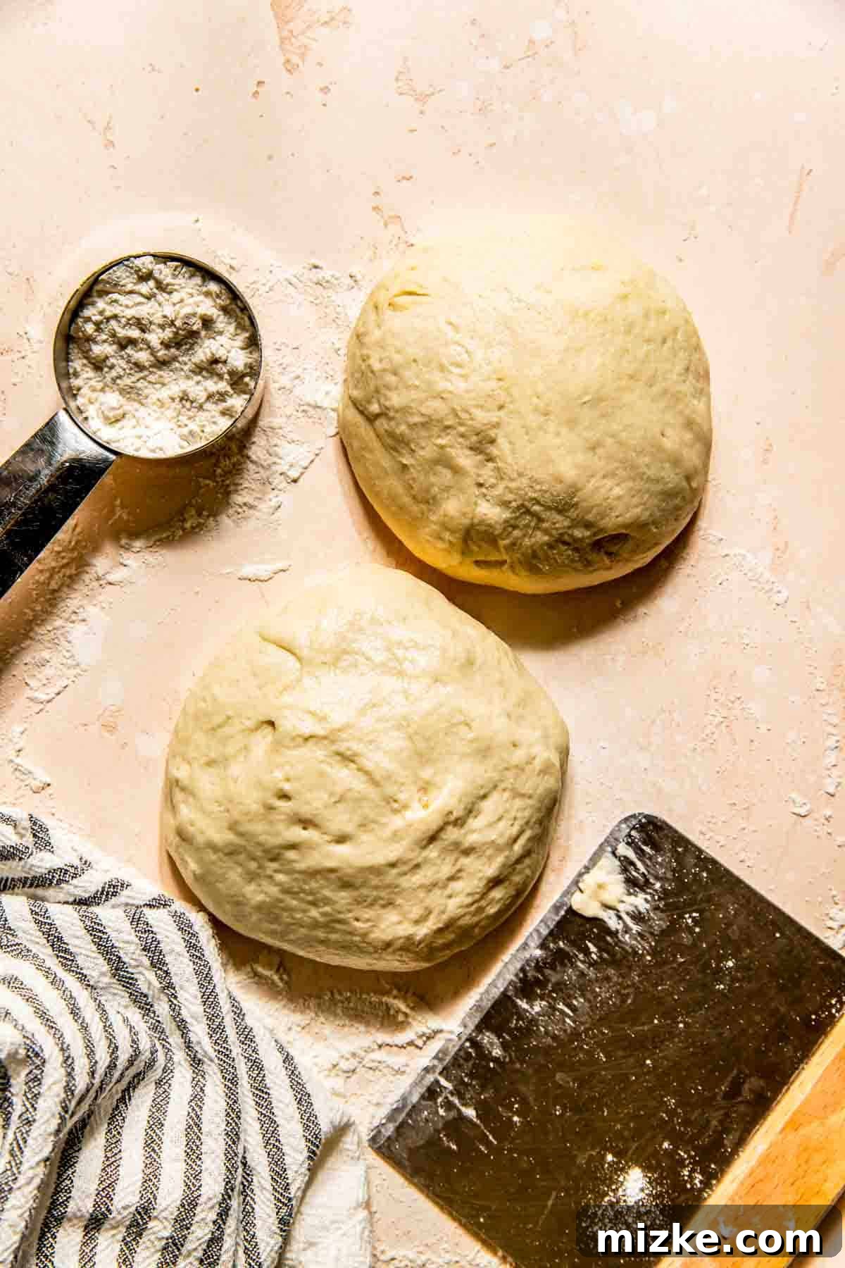 Crispy Thin Crust Pizza Dough Recipe 2 Two perfectly formed balls of thin crust pizza dough resting on a lightly floured surface, with a metal measuring cup of flour, a striped kitchen cloth, and a dough scraper nearby, ready for the baking adventure.