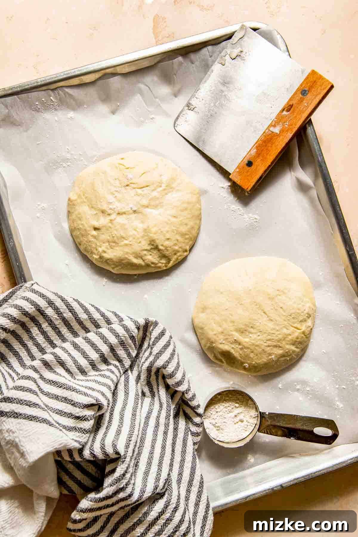 Crispy Thin Crust Pizza Dough Recipe 3 Two freshly prepared dough balls, intended for thin crust pizza, arranged neatly on a parchment-lined baking sheet, accompanied by a dough scraper, a striped kitchen towel, and a measuring cup holding flour.