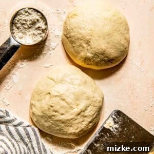 Crispy Thin Crust Pizza Dough Recipe 5 Two balls of dough on a floured surface with a metal measuring cup containing flour, a striped cloth, and a dough scraper nearby.