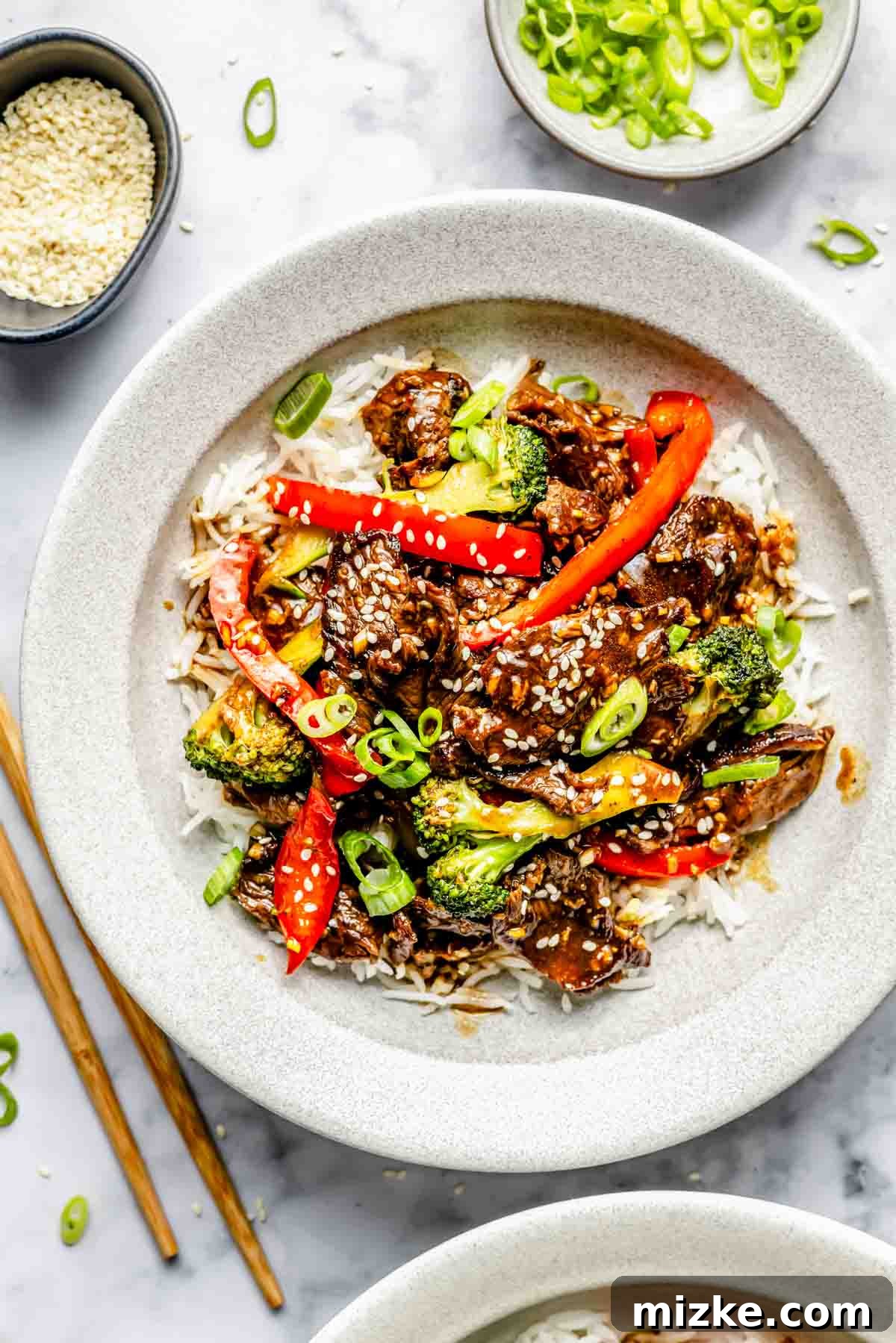 Quick-Slice Steak Stir-Fry 3 A bowl of rice topped with stir-fried beef, broccoli, red bell peppers, and sesame seeds, garnished with sliced green onions. Chopsticks lie beside the bowl.