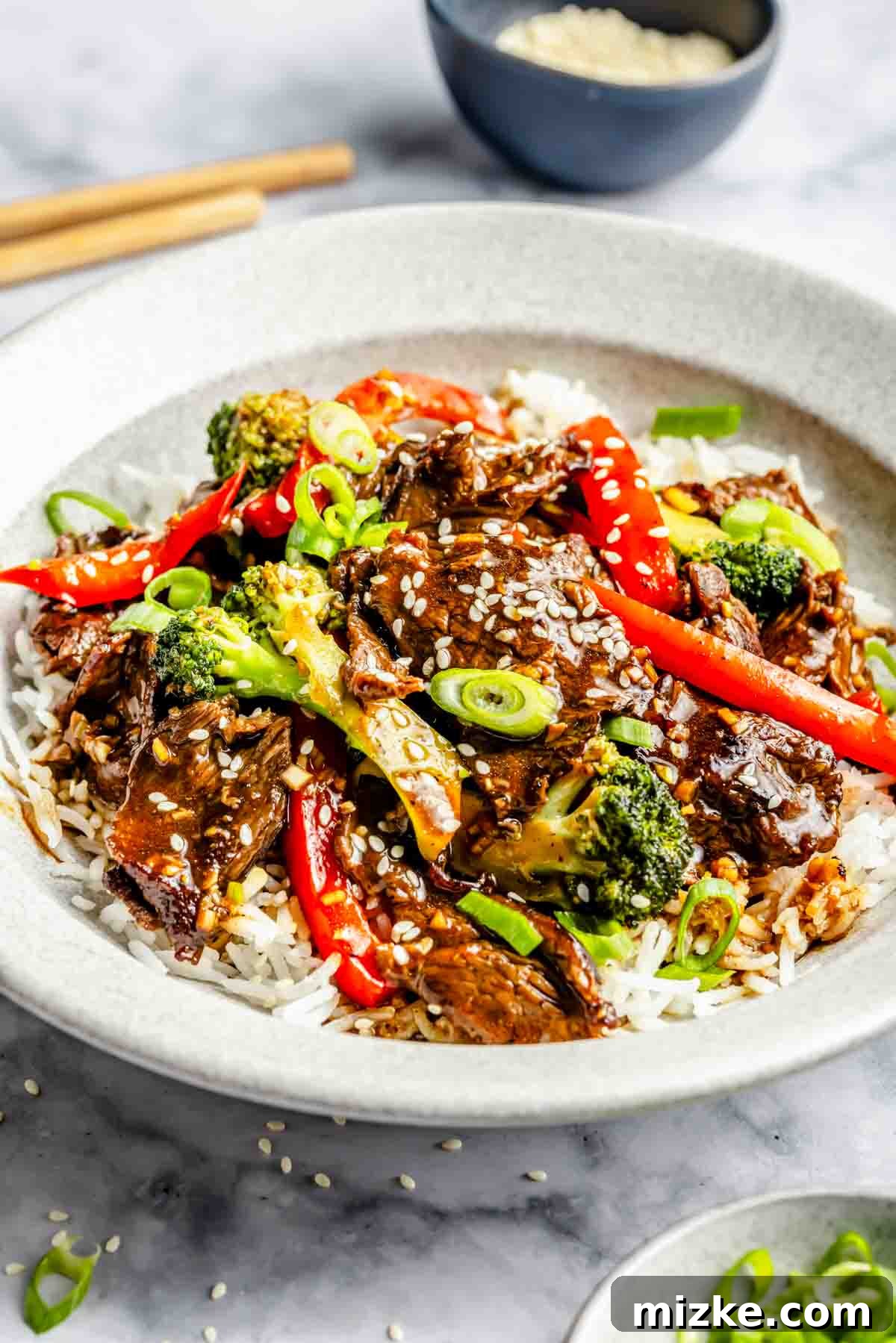 Quick-Slice Steak Stir-Fry 4 A bowl of rice topped with beef, broccoli, red bell peppers, and green onions, sprinkled with sesame seeds. A small bowl of sesame seeds is in the background.