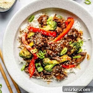 A bowl of rice topped with stir-fried beef, broccoli, red bell peppers, and sesame seeds, garnished with sliced green onions. Chopsticks lie beside the bowl.