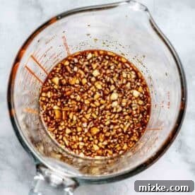 A glass measuring cup filled with a marinade containing chopped garlic and soy sauce, placed on a marble surface.