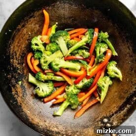 Stir-fried broccoli and red bell pepper slices in a large wok on a marble surface.