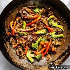 A wok filled with stir-fried beef slices, red bell peppers, and broccoli on a marble surface.
