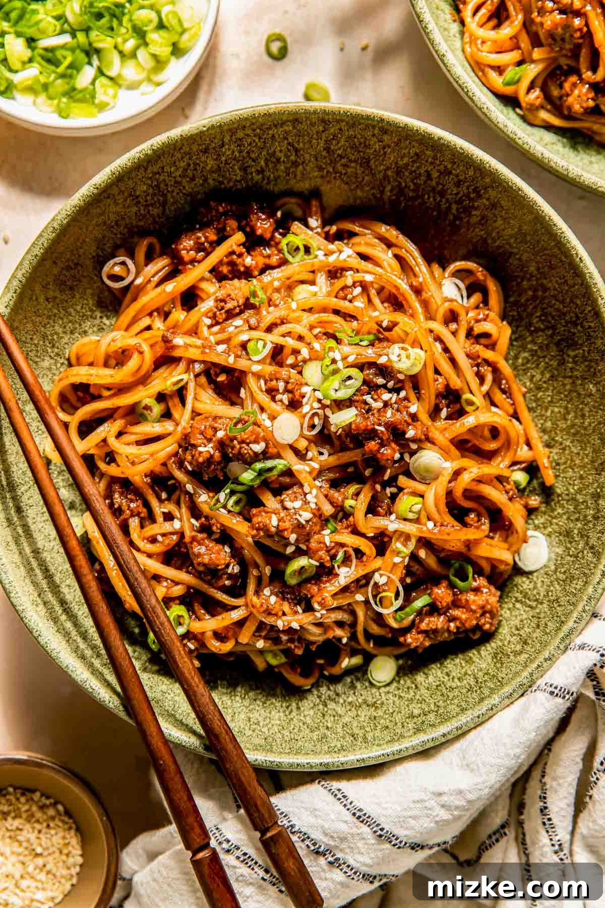 Sticky Mongolian Beef Noodles 3 A beautifully presented bowl of Mongolian ground beef noodles, garnished with fresh green onions and sesame seeds, with wooden chopsticks resting on the side.