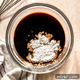 A glass bowl with soy sauce mixture and cornstarch is on a counter next to a metal whisk and striped cloth.