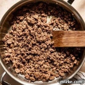 Cooked ground beef in a stainless steel pan with a wooden spatula, placed on a light-colored surface with a striped cloth nearby.