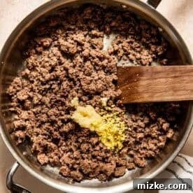 Ground beef cooking in a skillet with minced garlic and ginger, shown with a wooden spatula and a kitchen towel nearby.