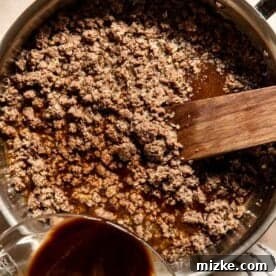 Ground beef browning in a skillet with a wooden spatula, next to a measuring cup containing sauce ingredients being poured in.