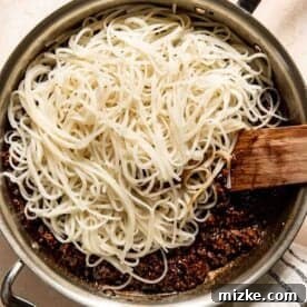 A pan with cooked rice noodles on top of a sauce mixture, stirred with a wooden spatula. A striped cloth lies beside the pan.