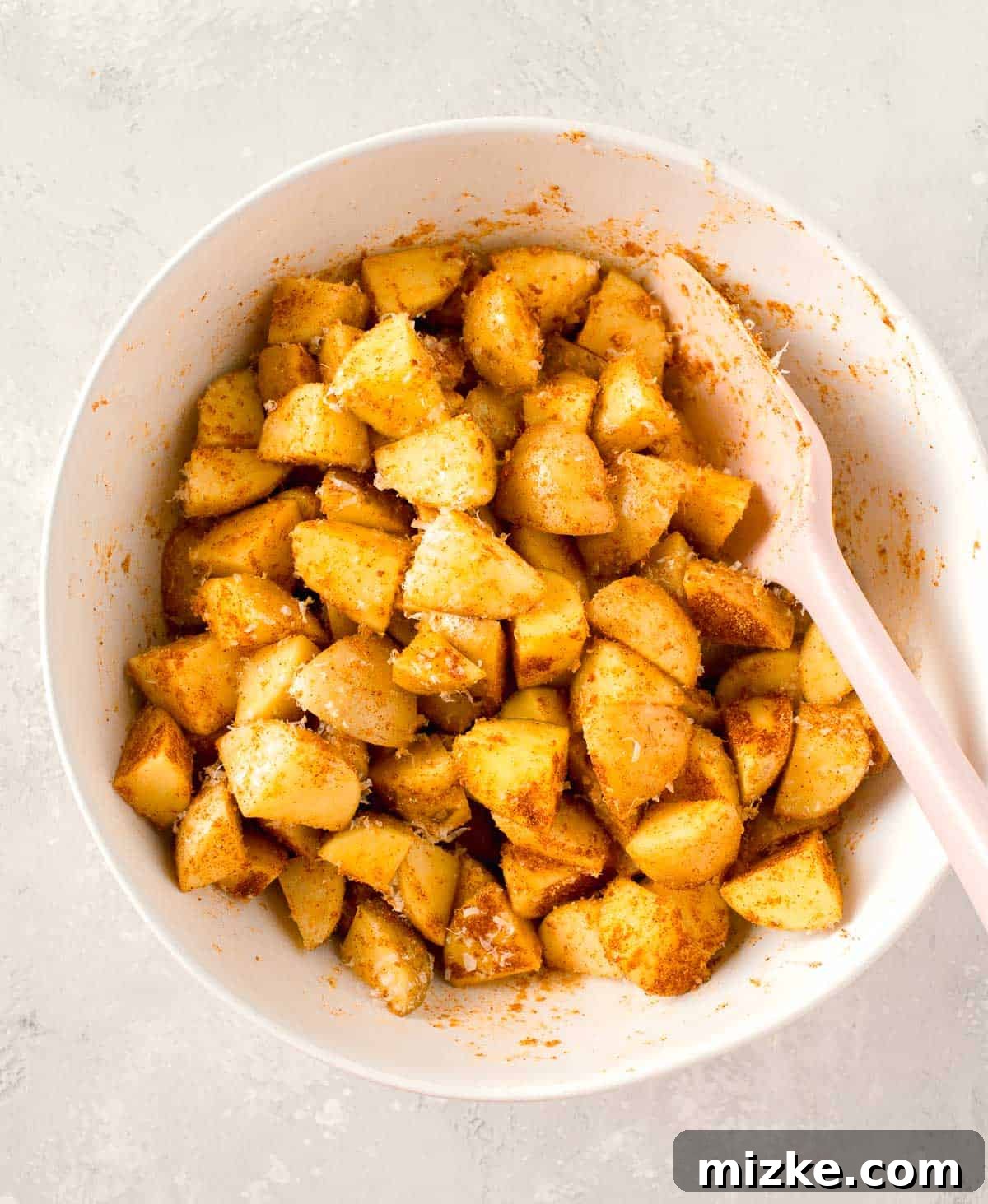 Crispy Garlic Parmesan Potatoes 5 seasonings and parmesan cheese mixed together with potatoes in a white bowl with pink silicone spatula