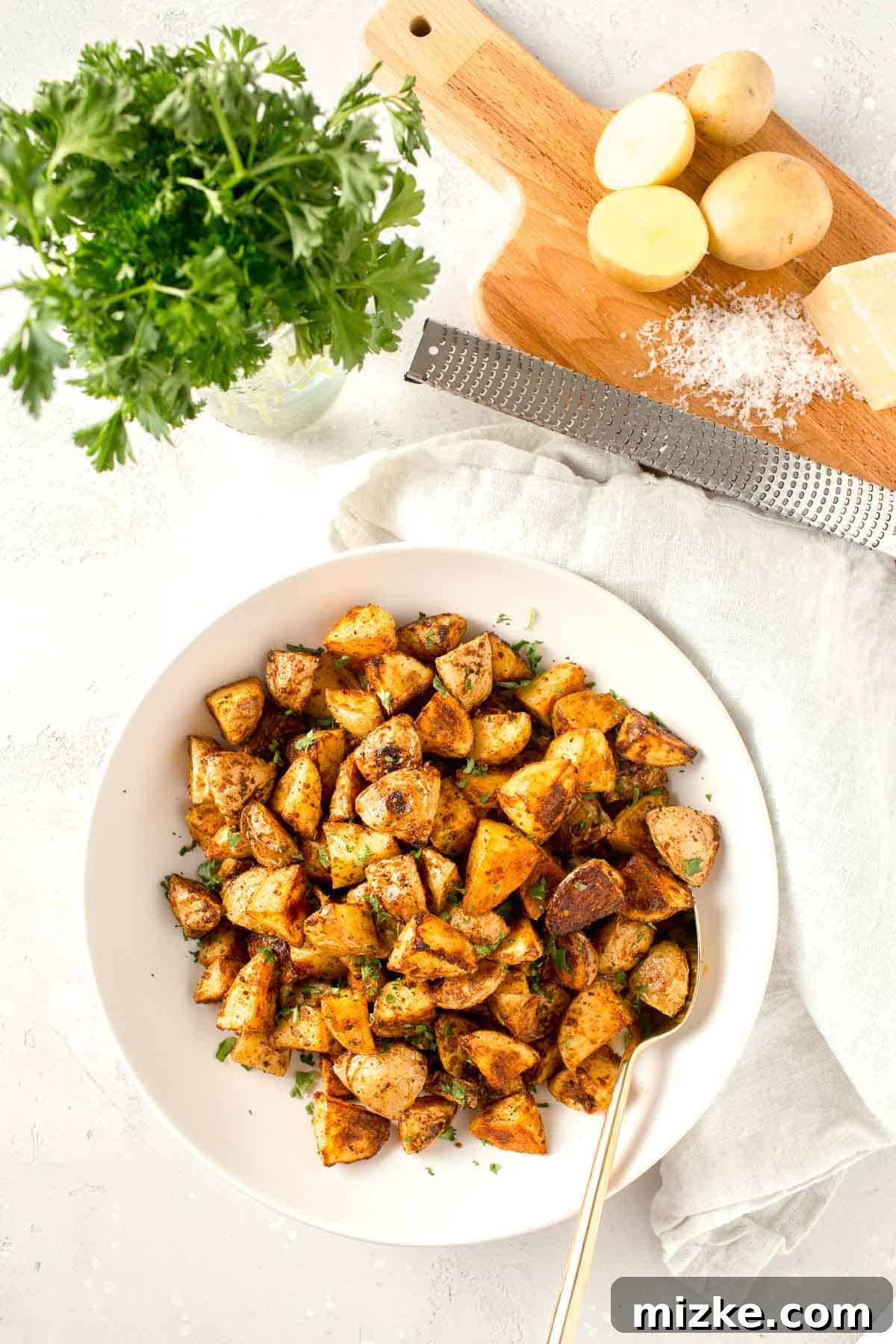 Crispy Garlic Parmesan Potatoes 8 overhead image of roasted parmesan potatoes in a large white bowl next to a linen towel, fresh parsley, metal zester, and freshly grated parmesan cheese