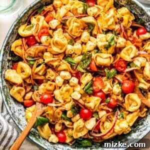 A bowl of tortellini pasta salad with cherry tomatoes, mozzarella balls, red onion, and fresh basil, mixed with a wooden spoon.