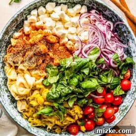Delicious Tortellini Pasta Salad 5 A large bowl filled with colorful ingredients for tortellini pasta salad: tortellini, cherry tomatoes, sliced red onion, mozzarella balls, fresh basil leaves, sliced pepperoncini, and assorted seasonings, with wooden utensils nearby for serving.