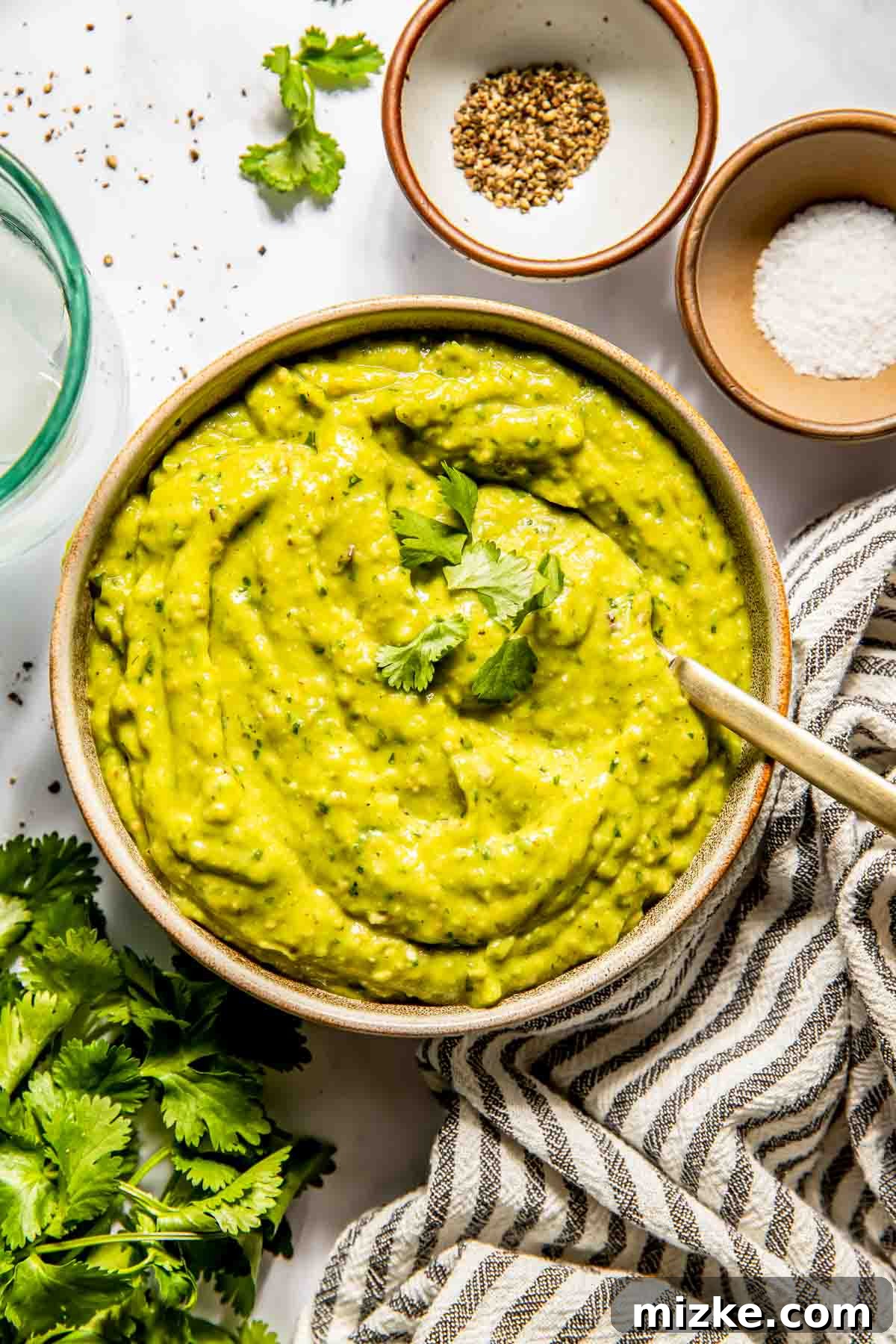 A beautifully presented bowl of vibrant tomatillo avocado salsa, garnished with fresh cilantro leaves, resting on a rustic striped towel. The scene is further enhanced by scattered cilantro sprigs, a glass jar, and small dishes containing aromatic spices and coarse salt, highlighting the fresh, natural ingredients used in this homemade salsa.