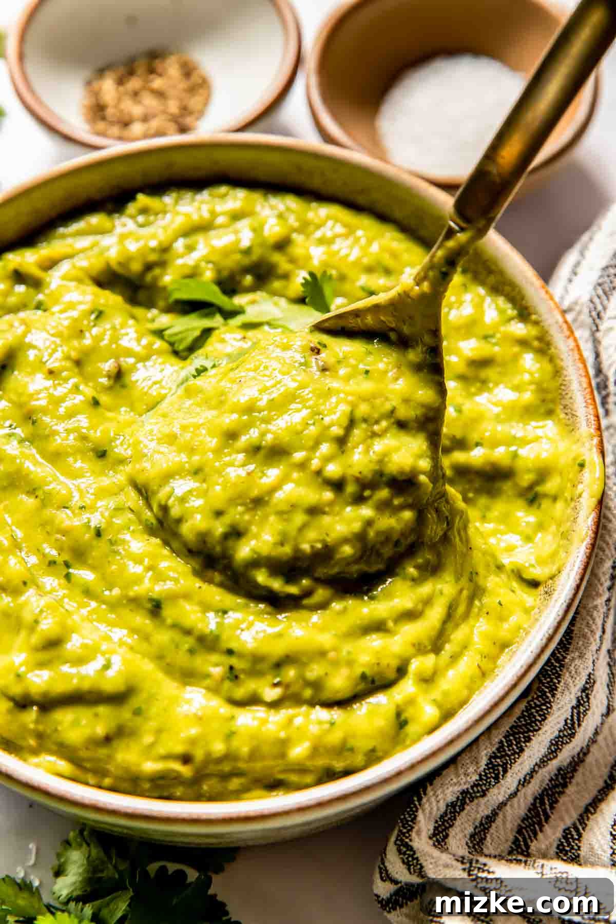 An overhead shot showcasing a bowl of exquisitely creamy tomatillo avocado salsa, with a spoon gently placed within the dip, indicating its readiness. The bowl is artfully arranged on a pristine white surface, surrounded by small, delicate bowls filled with an assortment of vibrant spices and a soft, striped linen cloth, emphasizing the freshness and culinary appeal of the dish.