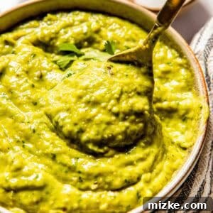 A bowl of creamy tomatillo avocado salsa with a spoon, surrounded by small bowls of spices and a striped cloth, showcasing the final dish prepared for serving.