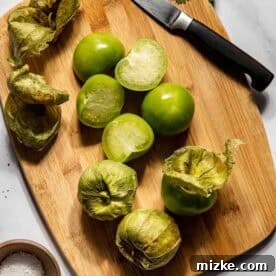 Several whole and halved tomatillos with husks removed, alongside a knife, salt, and pepper on a wooden cutting board with cilantro on a white surface, prepared for roasting.